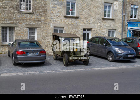 A US Army World War Two jeep parked between two modern cars in Sainte-Marie-du-Mont, Normandy, France. Stock Photo