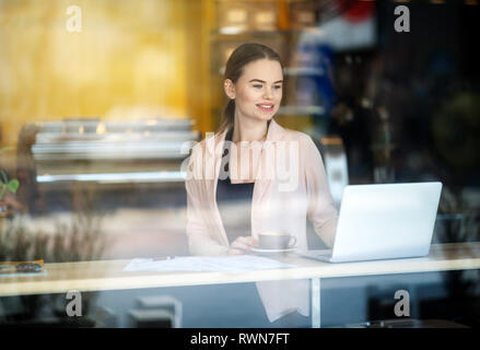 Smiling Woman working on laptop at coffee shop. Window Reflexions. Shot of a young woman using a laptop in a cafe Stock Photo