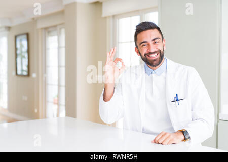 Handsome hispanic doctor or therapist man wearing medical coat at the clinic smiling positive doing ok sign with hand and fingers. Successful expressi Stock Photo