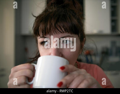 Young woman drinks coffee in the morning in the kitchen, tired eyes with red veins, close-up Stock Photo