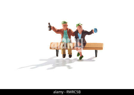 Miniature couple sitting on a bench celebrating toasting the camera saying cheers as they lift their beers in the air with shadow on white Stock Photo