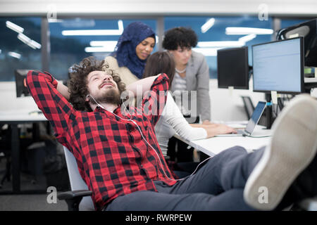young software developer resting with legs on desk while his multiethnics business team writing programming code in the background at startup office Stock Photo