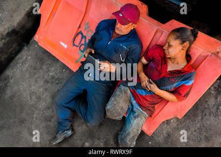 Colombian car mechanics read a mobile phone screen during a work break on the street in Barrio Triste, Medellín, Colombia. Stock Photo