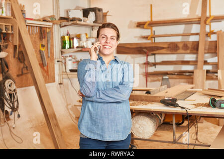 Young woman as a craftsman apprentice is talking on the mobile phone in the workshop Stock Photo
