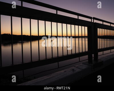 A sunset silhouette of a bridge over a river. Beautiful coloured sunset with striking silhouette of vertical lines from handrails looking to the river Stock Photo