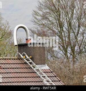 Chimney liner for log-burning stove curving from chimney pot during installation on a red tile roof. Roof ladder in place with tree background. Stock Photo