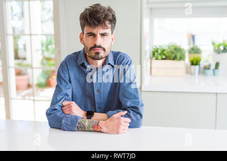 Young man wearing casual shirt sitting on white table skeptic and nervous, disapproving expression on face with crossed arms. Negative person. Stock Photo