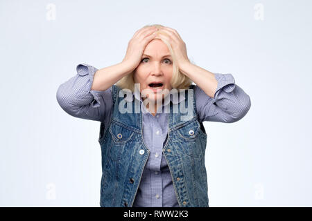 Anxious elderly blonde woman having frustrated stressed expression, holding hands on her head and looking in despair, being struck with panic because  Stock Photo