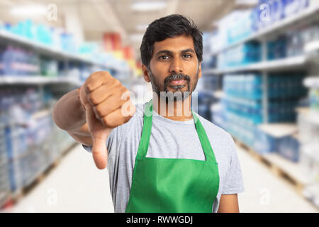 Indian male hypermarket or supermarket employee wearing green apron showing dislike gesture with thumb finger down and disappointed expression Stock Photo
