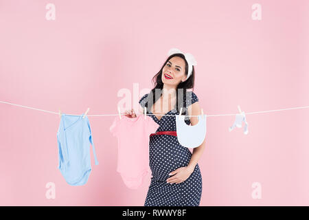 Pregnant woman in dotted dress hanging out baby clothes on clothesline on pink background Stock Photo