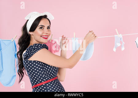 Excited pregnant woman hanging out baby clothes on clothesline isolated on pink Stock Photo