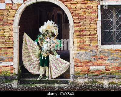 Venice, Italy - March 2, 2019 Person dressed in a typical Venetian Costume models under a arch towards a water channel in Venice during the Carnival o Stock Photo