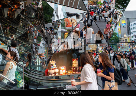 The glass shopping entrance to a mall in Harajuku, Tokyo, reflects people walking past in the street outside the building. Japan Stock Photo