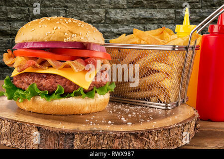 Cheeseburger with beef patty and bacon. French fries in basket, ketchup and mustard bottle in background. Isolated on brick wall. Real close up. Stock Photo