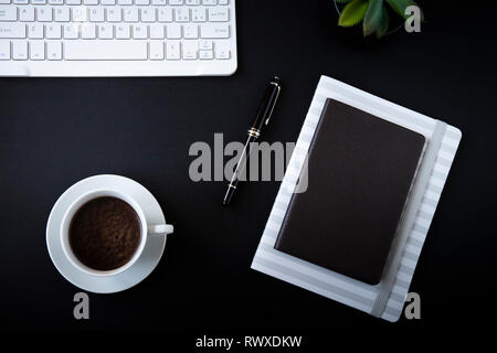 Office desk top view or flat lay with black background, white keyboard, cup of coffee, notebooks with black pen and a small succulent. Stock Photo