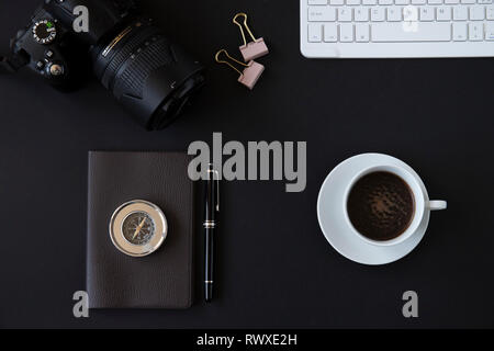 Office desk top view or flat lay with black background, a white keyboard, a cup of coffee, a small notebook with a black pen and a metal compass Stock Photo