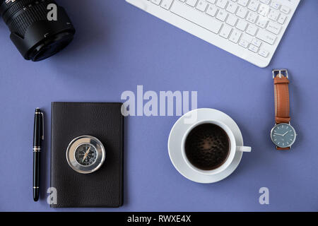 Office desk top view or flat lay with blue background, white keyboard, cup of coffee, notebook with black pen, leather watch, metal compass and camera Stock Photo