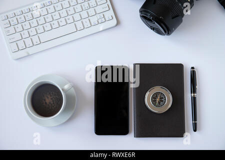 Office desk top view or flat lay with white background, white keyboard, reflex camera, cup of coffee, notebook with black pen, smartphone and a metal  Stock Photo