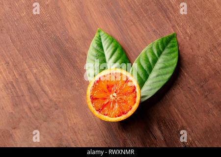 A closeup of an organic blood orange, shot from the top on a dark rustic wooden background with green leaves and a place for text Stock Photo