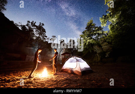 Young couple tourists man and woman resting beside camp and illuminated tent at night, warming hands by campfire. On background night starry sky full of stars and Milky way, mountain rocks, trees Stock Photo