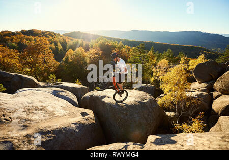 Professional cyclist riding on back wheel on trial bicycle, sportsman making acrobatic trick on the edge of big boulder on the top of mountain at sunset. Concept of extreme sport active lifestyle Stock Photo