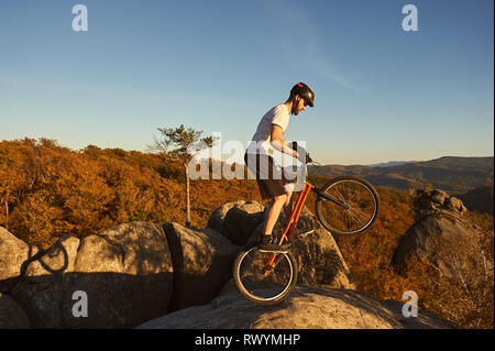 Professional cyclist balancing on back wheel on trial bicycle. Young sportsman making acrobatic stunt on the edge of big boulder on the top of mountain. Concept of extreme sport active lifestyle Stock Photo