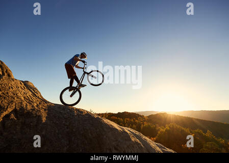 Silhouette of professional cyclist riding on back wheel on trial bike. Fearless sportsman biker making acrobatic stunt on the edge of big boulder at sunset. Concept of extreme sport active lifestyle Stock Photo