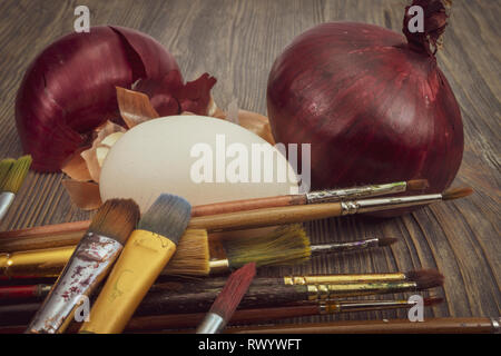 Fresh egg, painting brushes, whole red onion and dried onion skin on a wooden table. Making Easter eggs dyed with onion skins concept Stock Photo