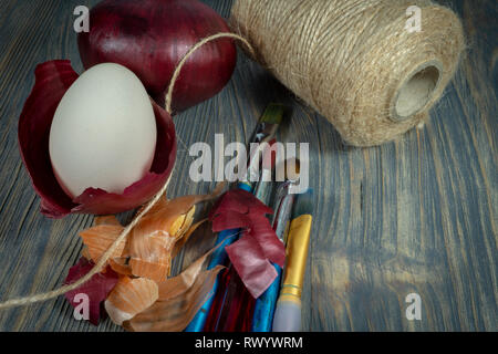 Fresh egg balancing in a dried onion skin, painting brushes, ball of brown twine and dried onion skin on a wooden table. Easter eggs dyed with onion s Stock Photo