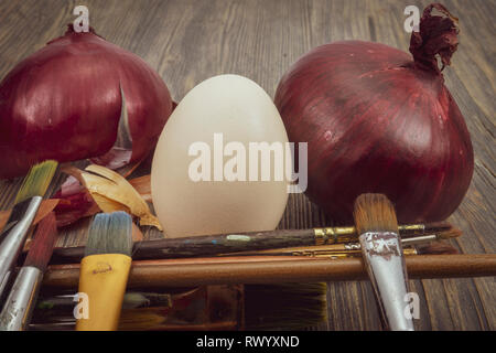 Fresh whole red onion, egg and dried onion skin, painting brushes on a wooden table. Making Easter eggs dyed with onion skins concept Stock Photo