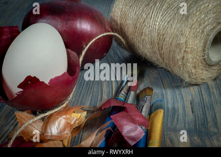 Fresh whole red onion, egg balancing in a dried onion skin and ball of brown twine on a wooden table. Making Easter eggs dyed with onion skins concept Stock Photo