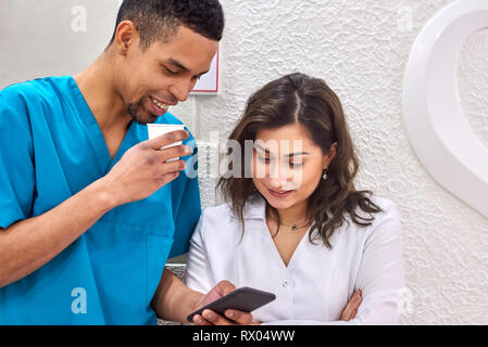 Young male african and female caucasian dentists on coffee break. Smiling and showing stuff on the smartphone Stock Photo