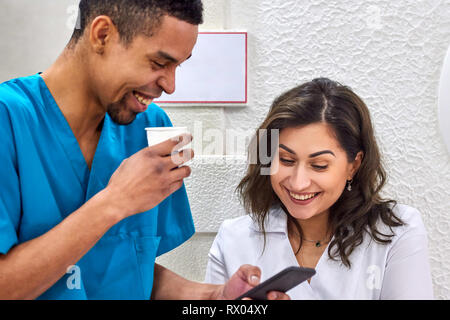 Young male african and female caucasian dentists on coffee break. Smiling and showing stuff on the smartphone Stock Photo