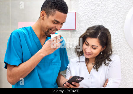 Young male african and female caucasian dentists on coffee break. Smiling and showing stuff on the smartphone Stock Photo