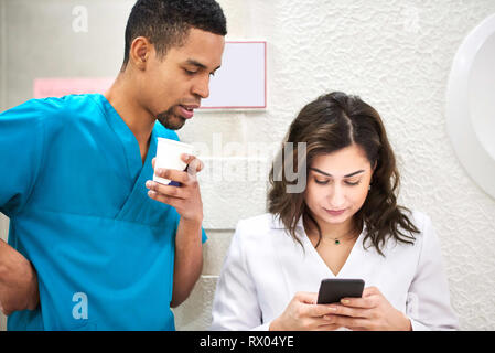 Young male african and female caucasian dentists on coffee break. Smiling and showing stuff on the smartphone Stock Photo