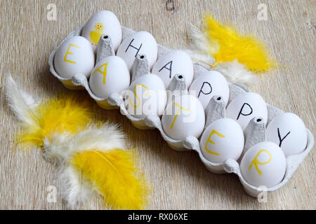 Happy Easter written on white eggs in a box and yellow feathers on wooden background Stock Photo