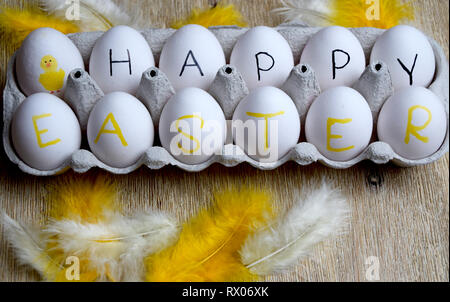 Happy Easter greetings on white eggs in a box and yellow feathers on wooden background Stock Photo