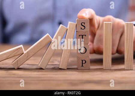 Businessperson's Finger Over Wooden Blocks With Stop Text Stopping Falling Dominos On Wooden Desk Stock Photo