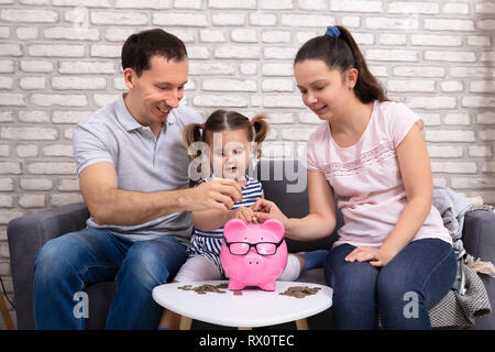 Happy Parents With There Daughter Inserting Coin In Pink Piggy Bank Stock Photo