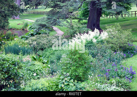 Gravel path through lush english garden with cottage flowers, shrubs, old trees . Stock Photo