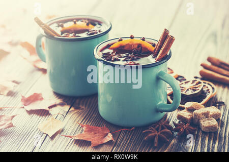 Mulled wine in blue enameled rustic mugs with spices and citrus fruits on wooden table with autumn leaves. Retro toned photo. Stock Photo