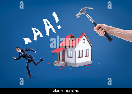 A hand holding a hammer breaking small house's roof with a businessman running away screaming. Stock Photo