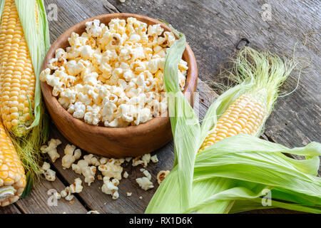 Prepared popcorn in wooden bowl, corncobs on wooden table. Stock Photo