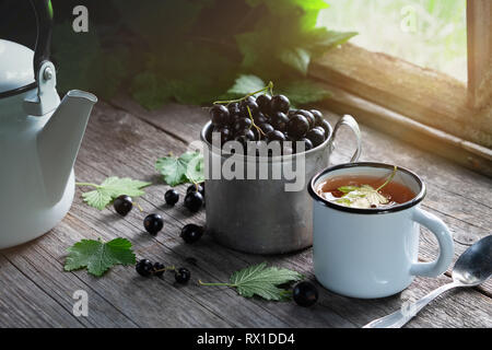 Mug of black currants, enameled cup of fruit tea with black currant berries, tea kettle on wooden table in a retro village house. Stock Photo