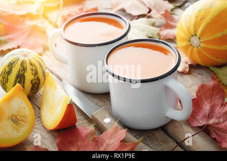 Two white enameled cups of pumpkin juice, pumpkins and fallen maple leaves on table. Stock Photo