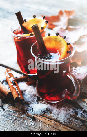 Glass mugs of mulled wine with spices and citrus fruits on wooden table with autumn leaves and snow. Stock Photo
