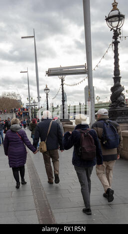 Two couples of senior citizens, warmly dressed, walking on the South Bank, London, UK towards Westminster Bridge on a cloudy and blustery day in March Stock Photo