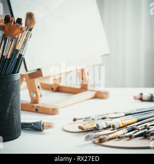 Artistic equipment in painter studio: easel, paint brushes, tubes of paint, palette and paintings on work table of artist. Stock Photo