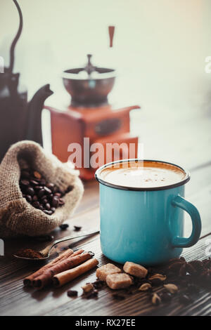 Espresso coffee in blue enameled cup, sugar cubes, sack of coffee beans, spices, coffee mill on wooden table. Retro toned. Stock Photo