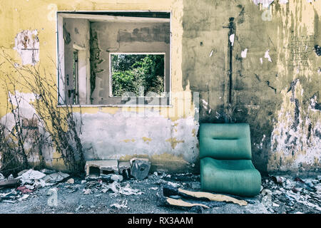 Grungy wallpaper background of yellow abandoned construction with windows showing nature and a green worn sofa in front of it. No people. Stock Photo